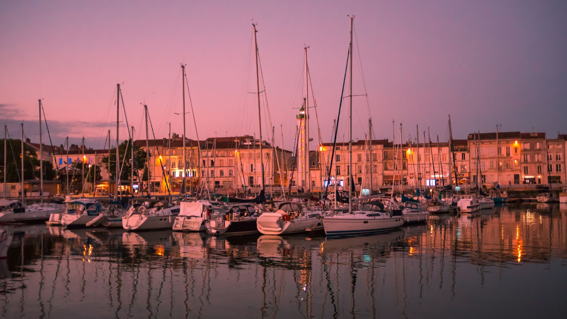 Vue sur les voiliers amarrés au Vieux-Port de La Rochelle, leurs mâts se dressant face aux façades historiques illuminées du port. L’eau reflète les lumières des immeubles anciens, offrant une ambiance paisible et typiquement rochelaise au coucher du soleil.
